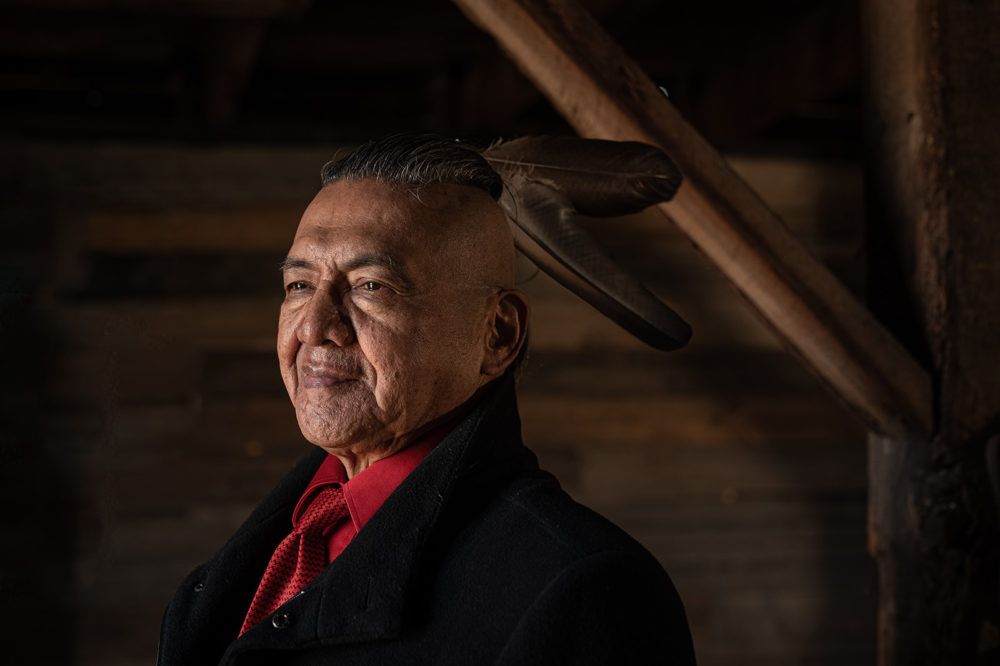 ageing native man with feathers tied in his hair wearing a black suit with red shirt and tie poses for sechelt headshots with sherry nelsen photography